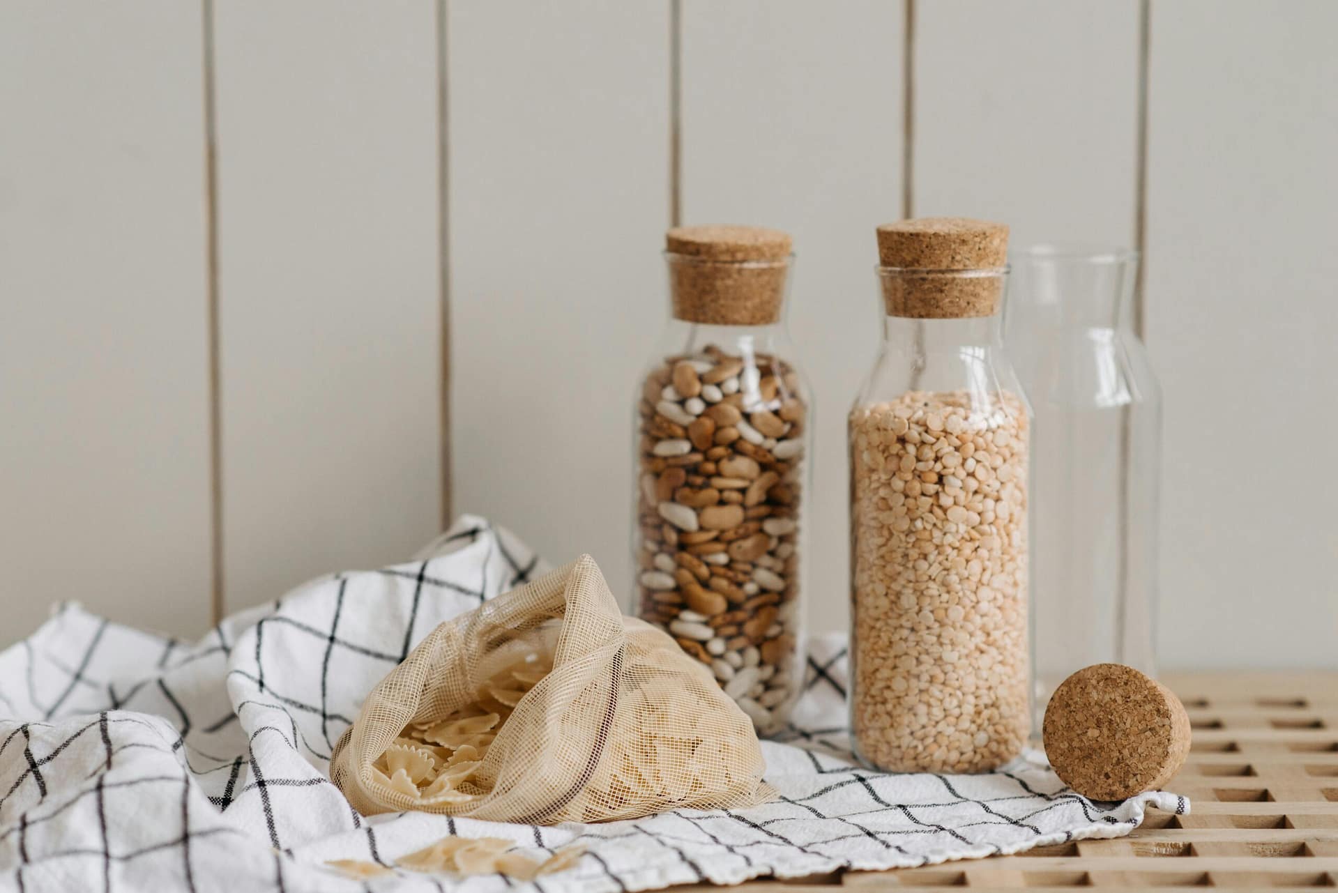 Eco-friendly jars filled with beans and grains, featuring a mesh bag and cloth on a wooden surface.